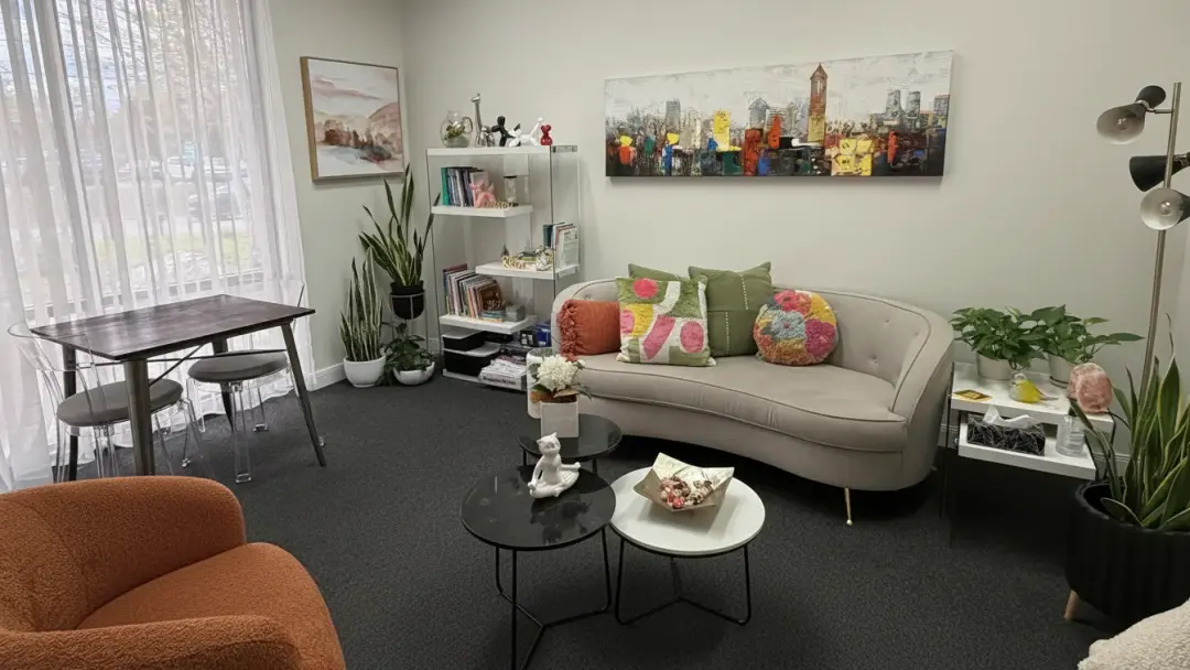 The warm Oakville therapy office: a cream sofa with colourful pillows under a city-skyline painting, plants and a soft-curtained window